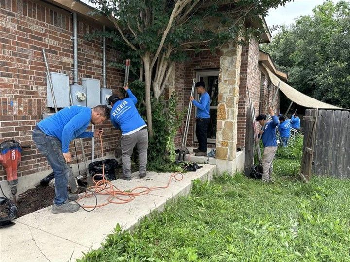 Workers in blue shirts install utilities outside a brick building with overgrown vegetation. Workers in blue shirts install utilities outside a brick building with overgrown vegetation.