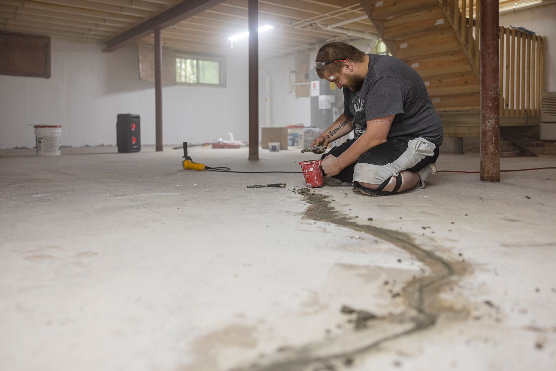 Worker sealing concrete floor cracks during foundation repair in a basement with construction tools.