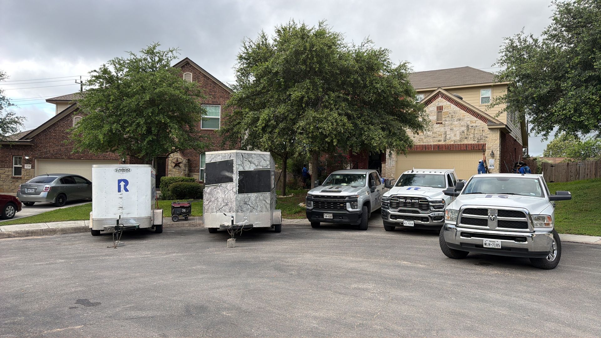 White trucks and utility vehicles parked in a residential driveway with two trees in front of houses