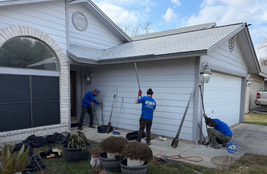 Three workers in blue shirts use long-handled tools to clean the exterior siding of a suburban house. Three workers in blue shirts use long-handled tools to clean the exterior siding of a suburban house.