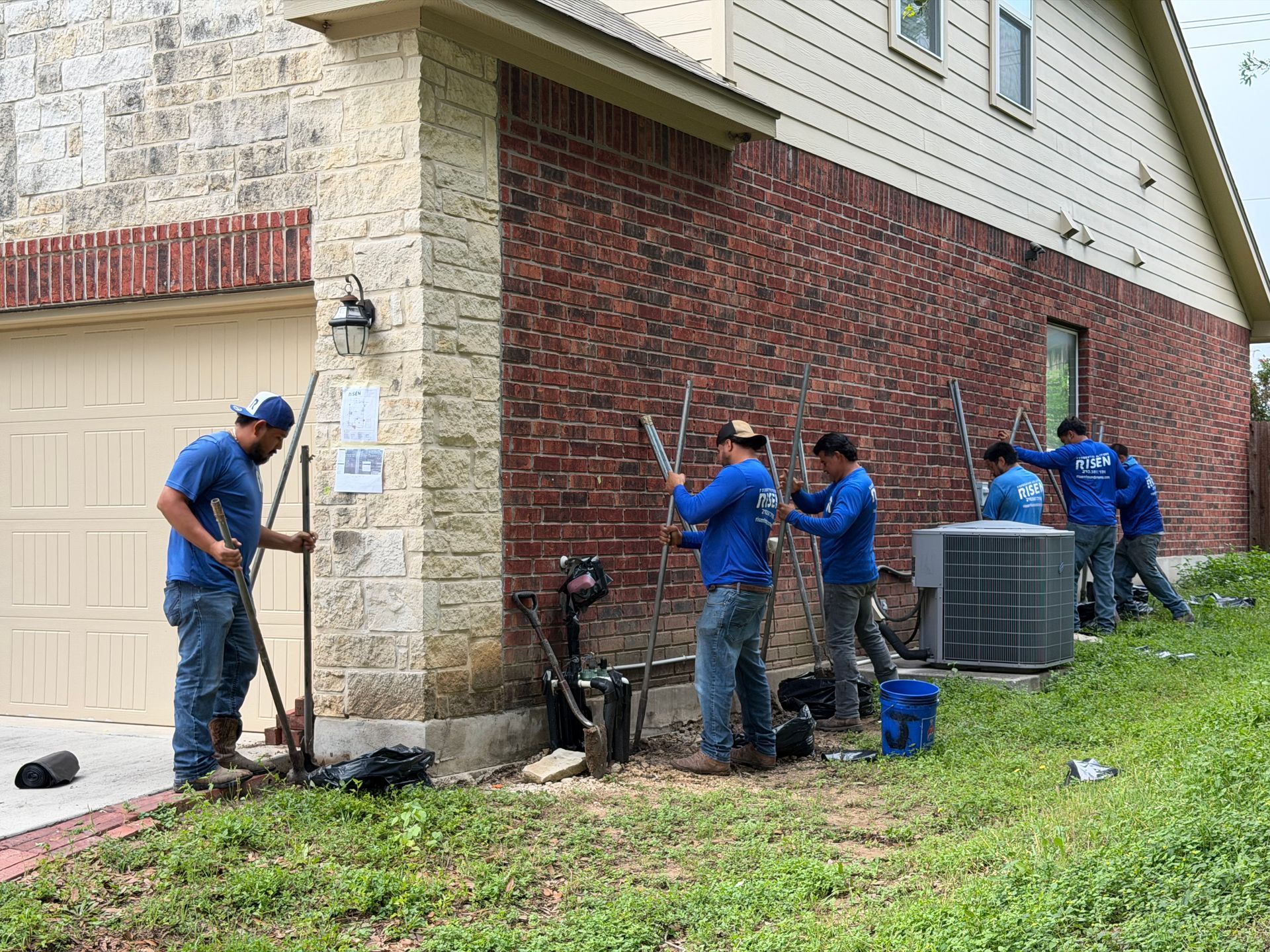 Workers repair an outdoor brick wall beside a house, with tools and an air conditioner unit nearby Workers repair an outdoor brick wall beside a house, with tools and an air conditioner unit nearby