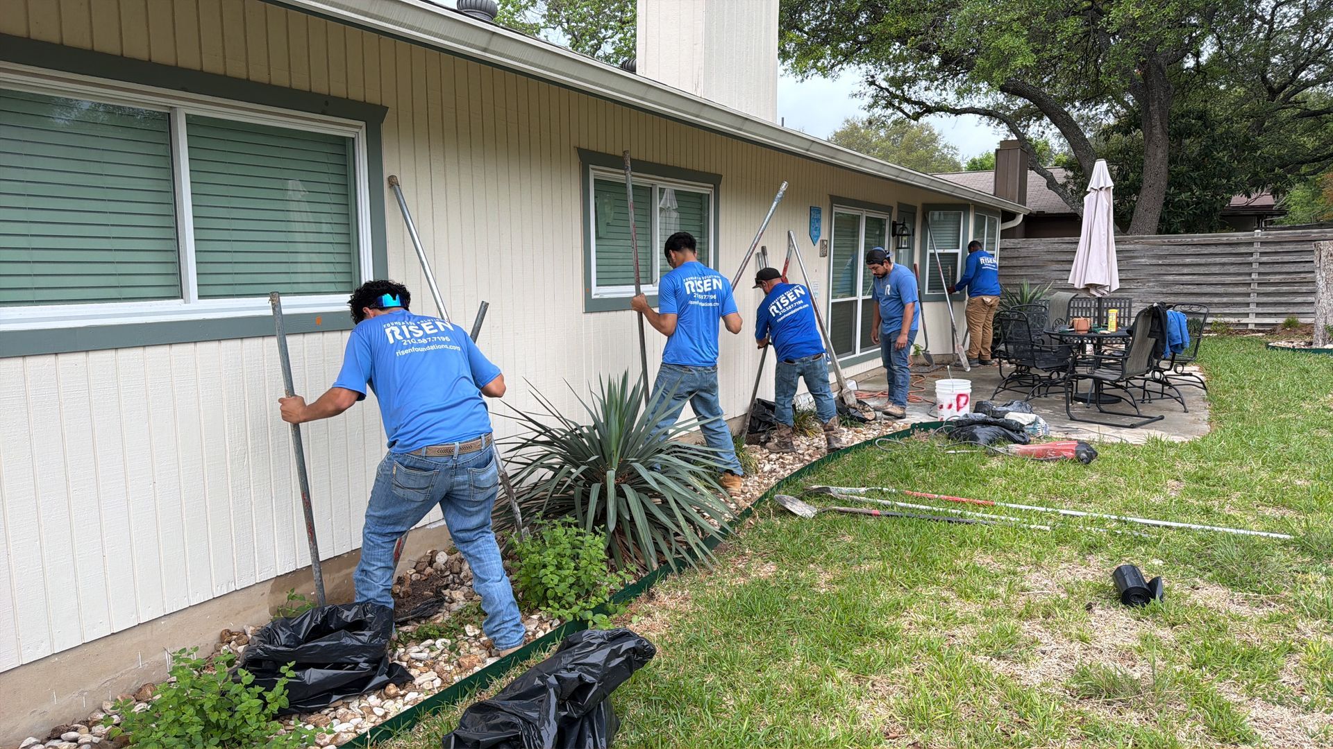 Workers landscaping along the side of a beige house, planting and trimming garden beds.