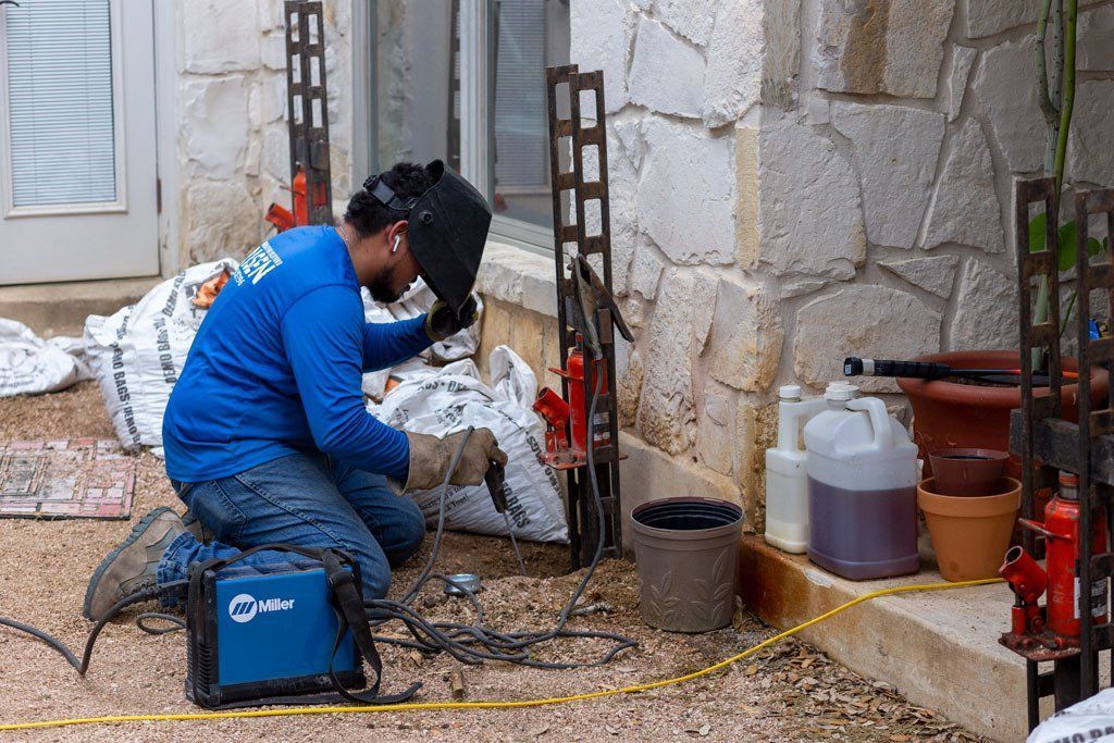 Worker in blue welding gear crouches beside stone wall, welding metal outdoors with tools and gas canisters nearby