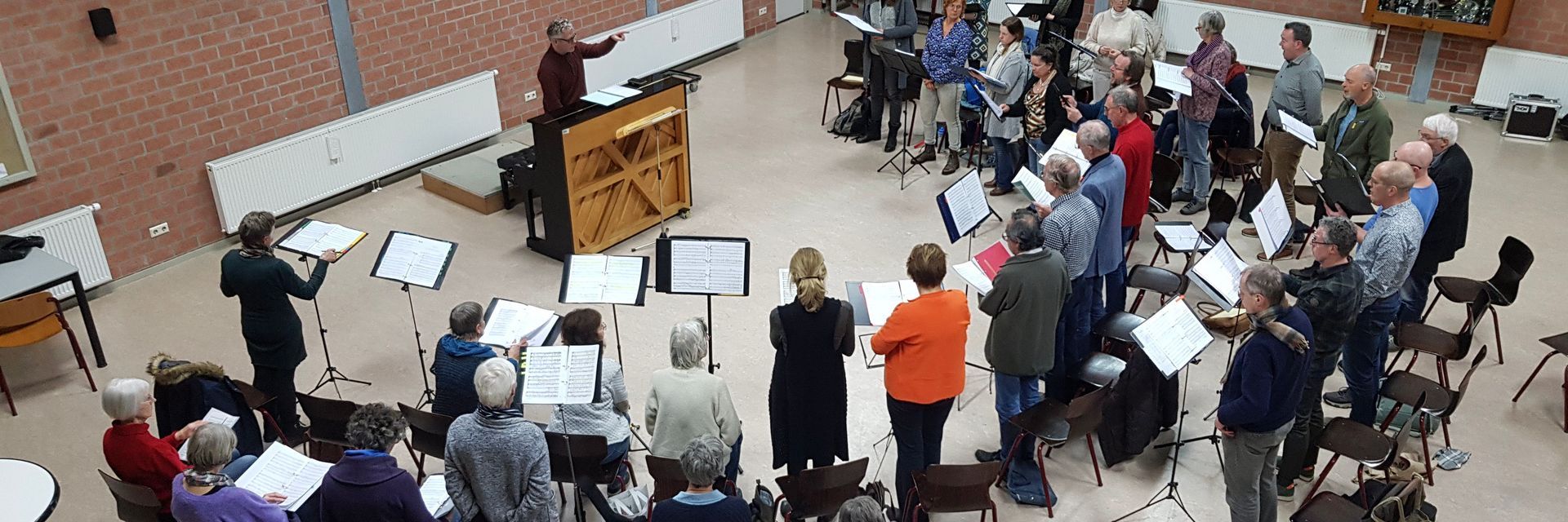 Een koor repeteert in een grote zaal met een dirigent achter de piano. De leden houden bladmuziek vast.