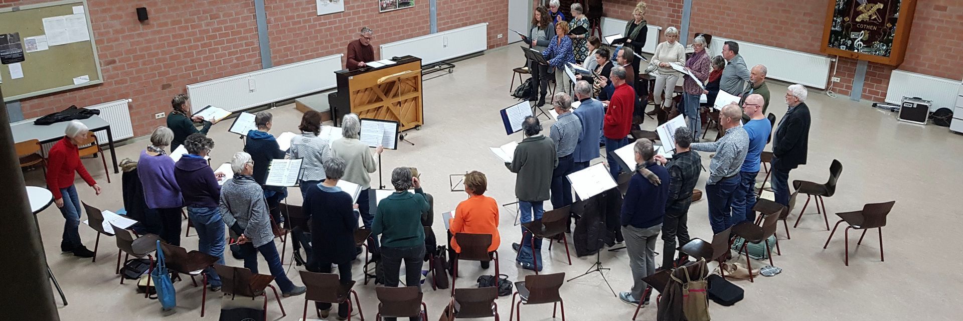 Een koor repeteert in een grote zaal. Mensen staan ​​met bladmuziek in hun handen. In het midden staat een piano.