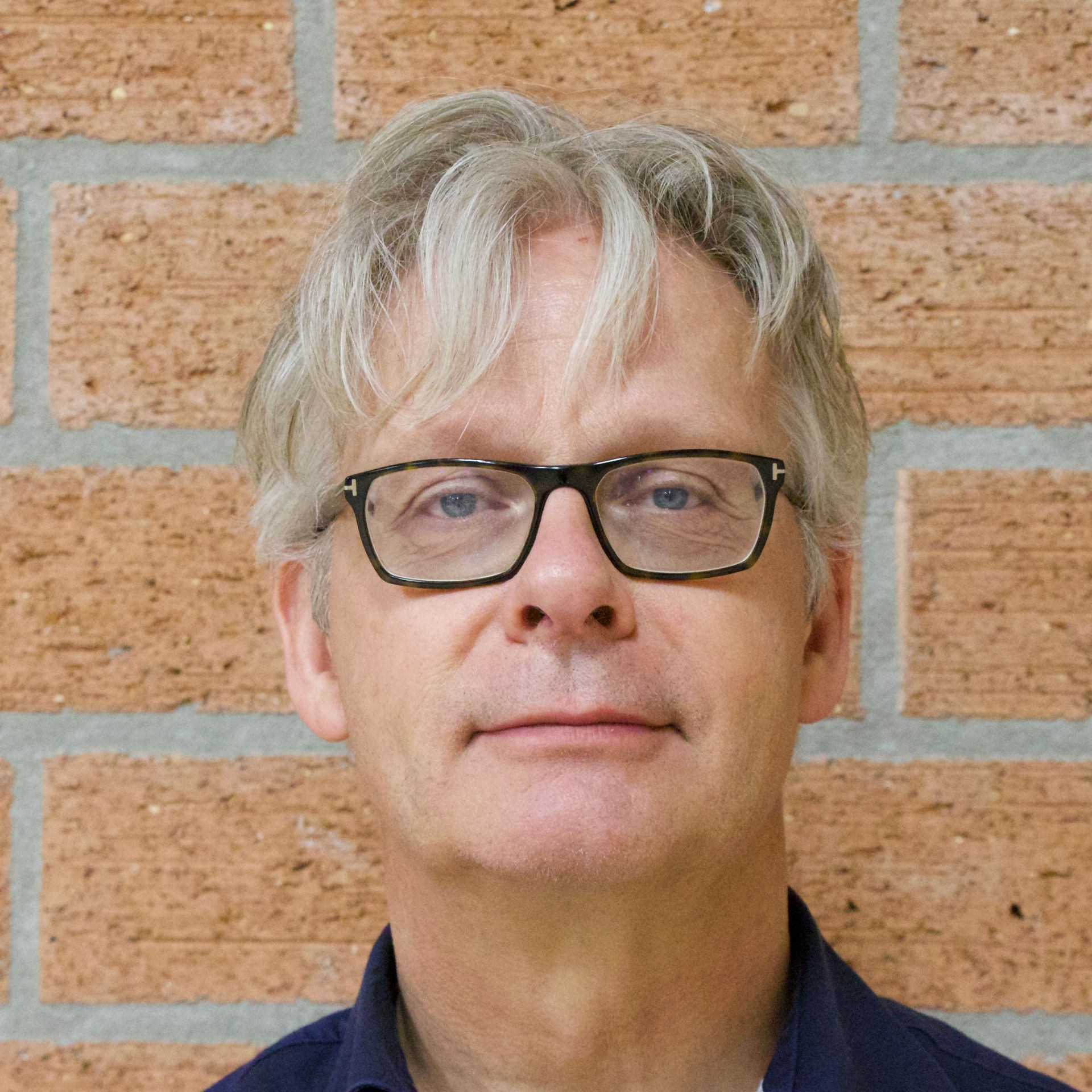 Man met bril en grijs haar tegen een bakstenen muur.