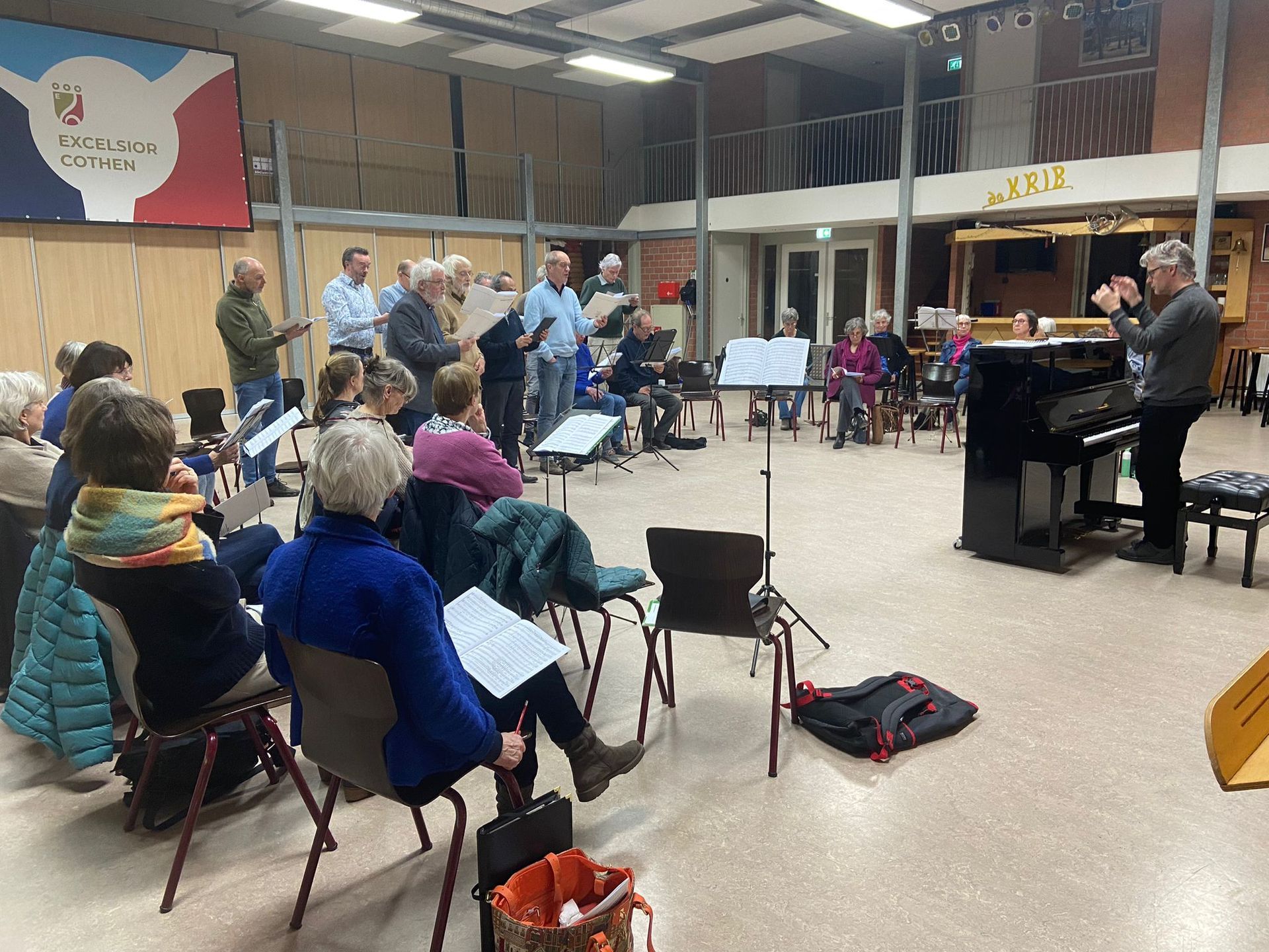 Een koor repeteert in een felverlichte zaal, met een pianist, lessenaars en zangers die naar bladmuziek kijken.