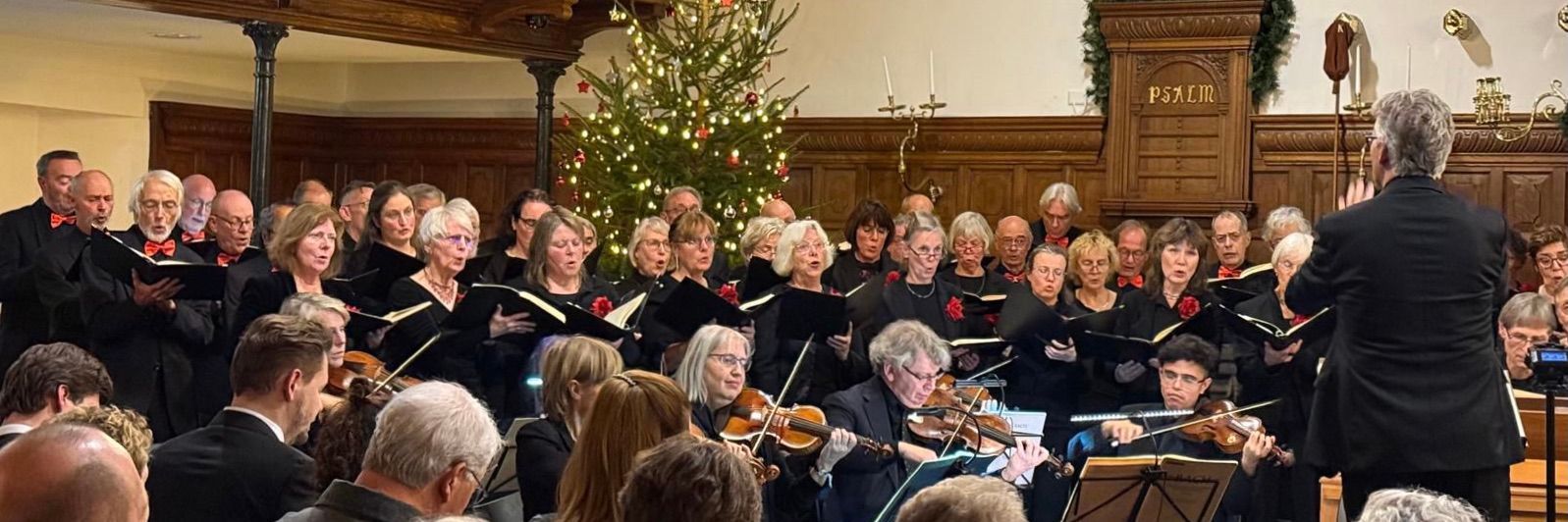 Koor en orkest treden op in een zaal met houten lambrisering, de dirigent staat vooraan, een kerstboom op de achtergrond.