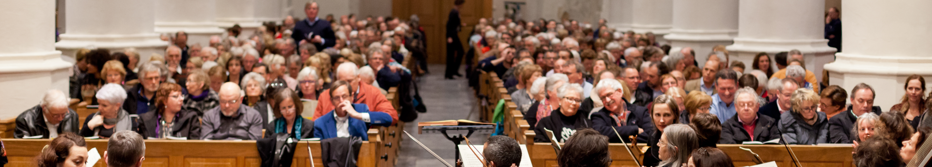 Publiek in een grote zaal, waarschijnlijk een conferentie- of lezingzaal. Mensen zitten in rijen naar voren gericht.