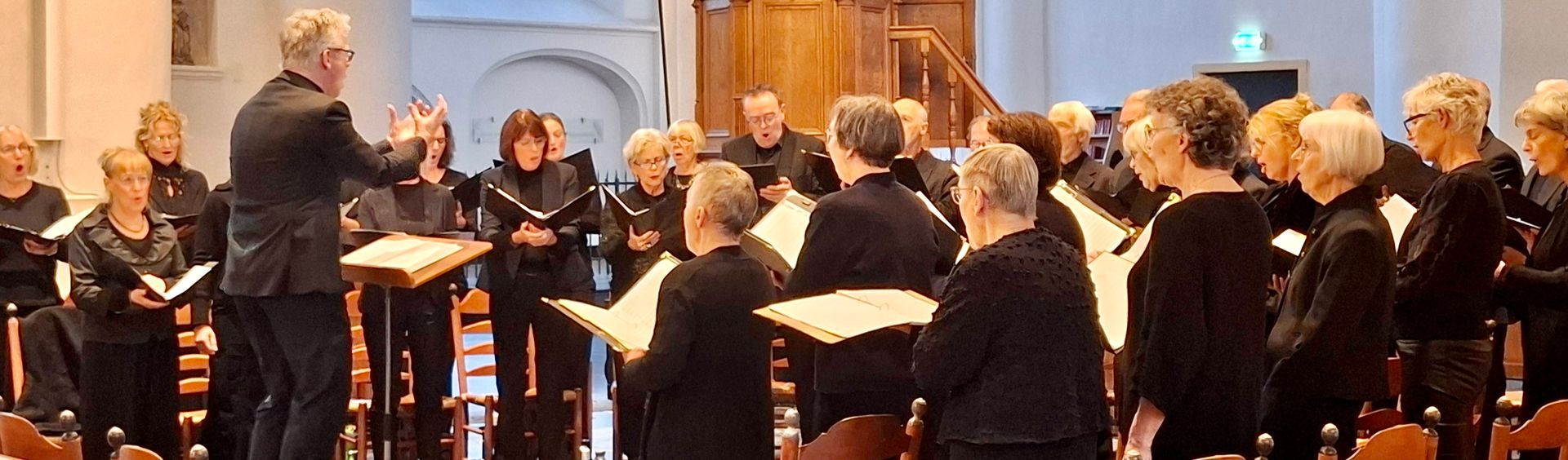 Een koor in een kerk, de dirigent staat tegenover de groep. De zangers houden bladmuziek vast en dragen zwarte gewaden.
