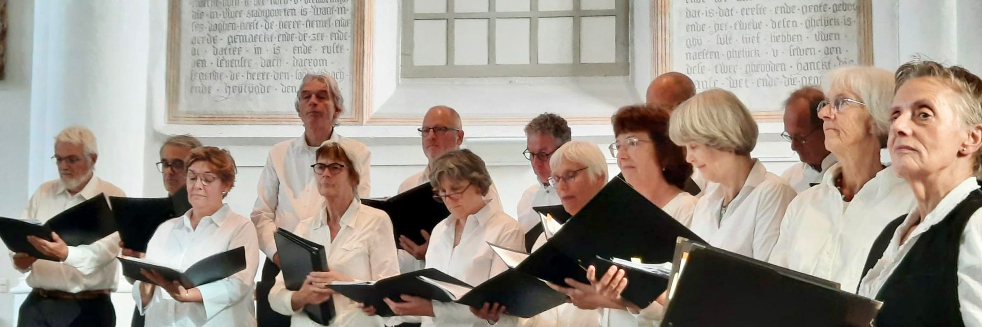 Een koor van ouderen zingt in een witgekalkte ruimte met teksten op de muren, terwijl ze bladmuziek vasthouden.