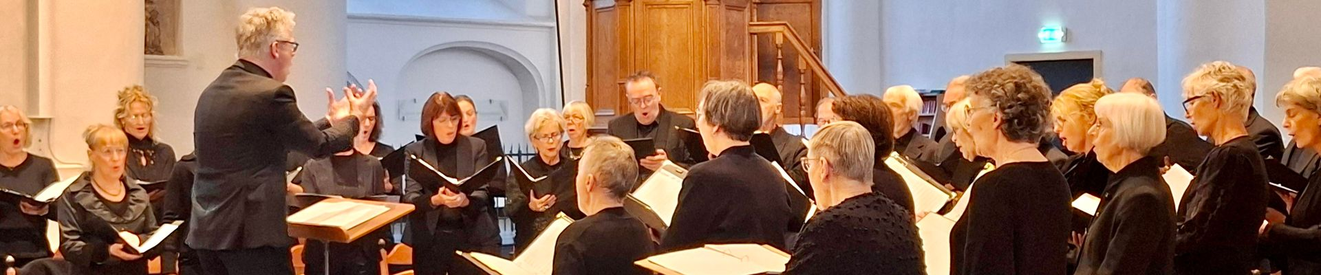 Een koor treedt op in een zaal; de dirigent maakt gebaren, de zangers lezen de muziek en dragen zwarte gewaden.