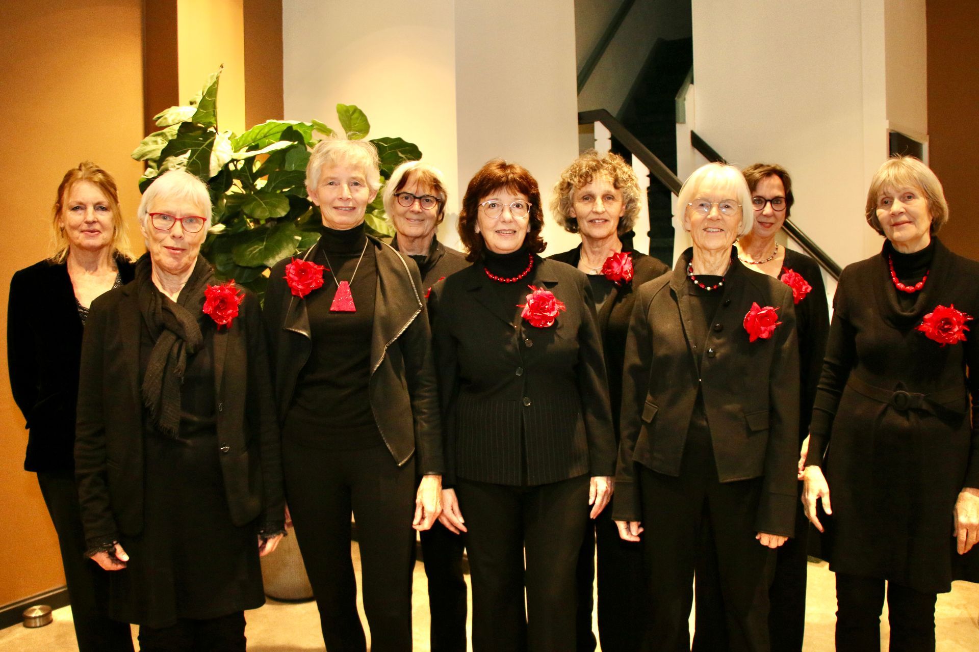 Een koorgroep in zwarte outfits met rode bloemspelden poseert binnen bij een plant.