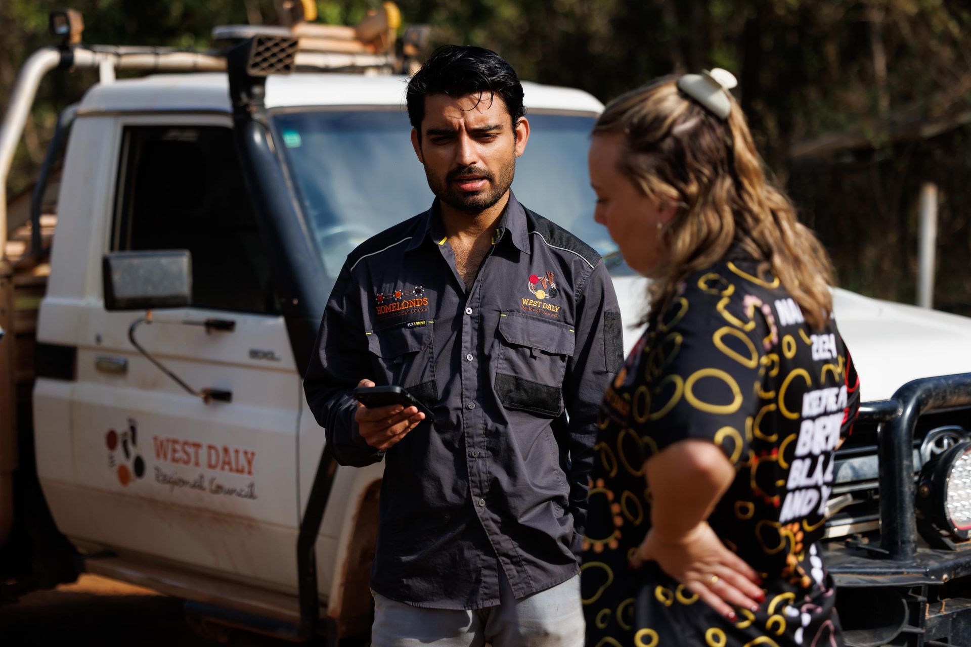 Man in gray shirt showing phone to woman near a white truck, outdoors.