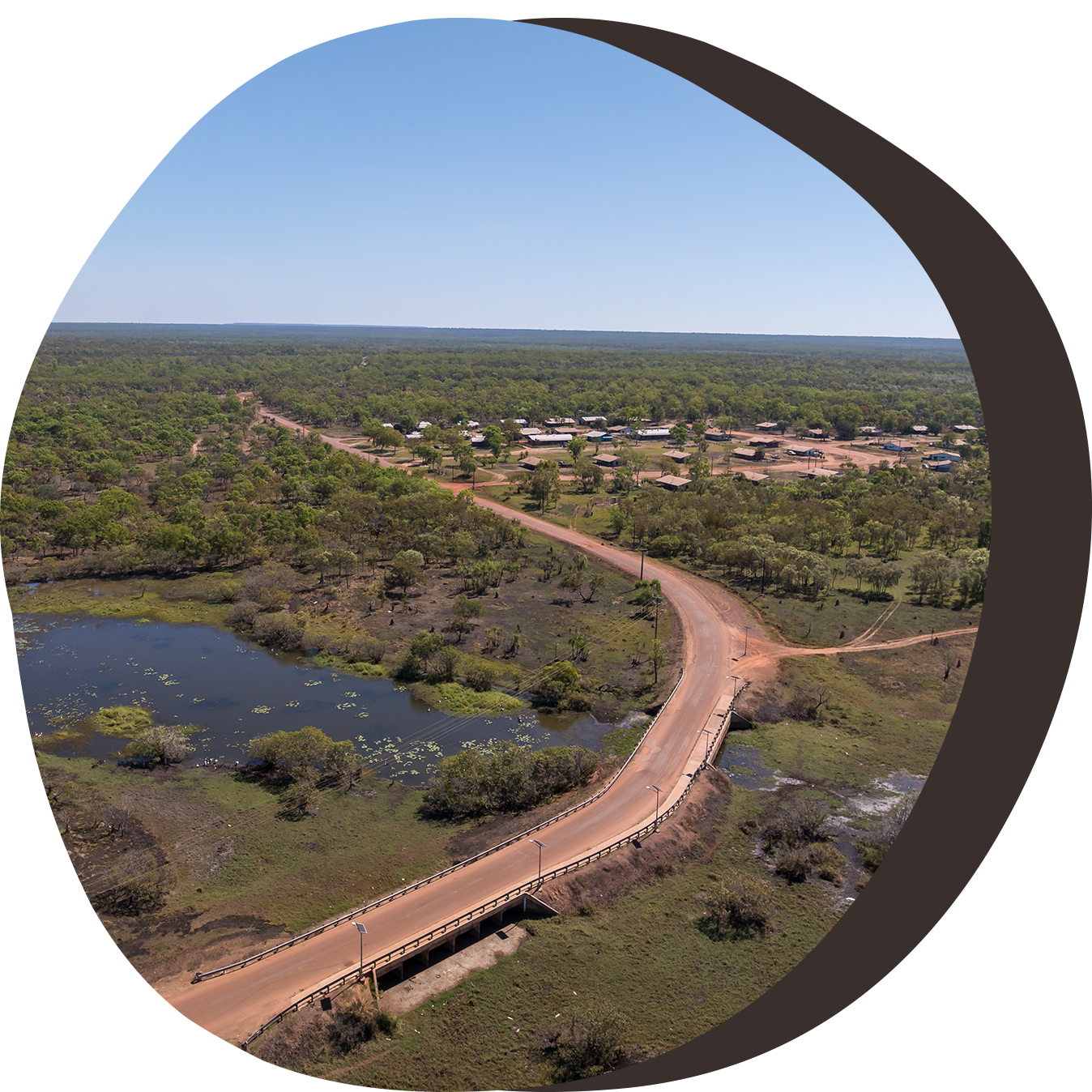 Aerial view: road bridge over water, leading to a small settlement surrounded by green trees under a blue sky.