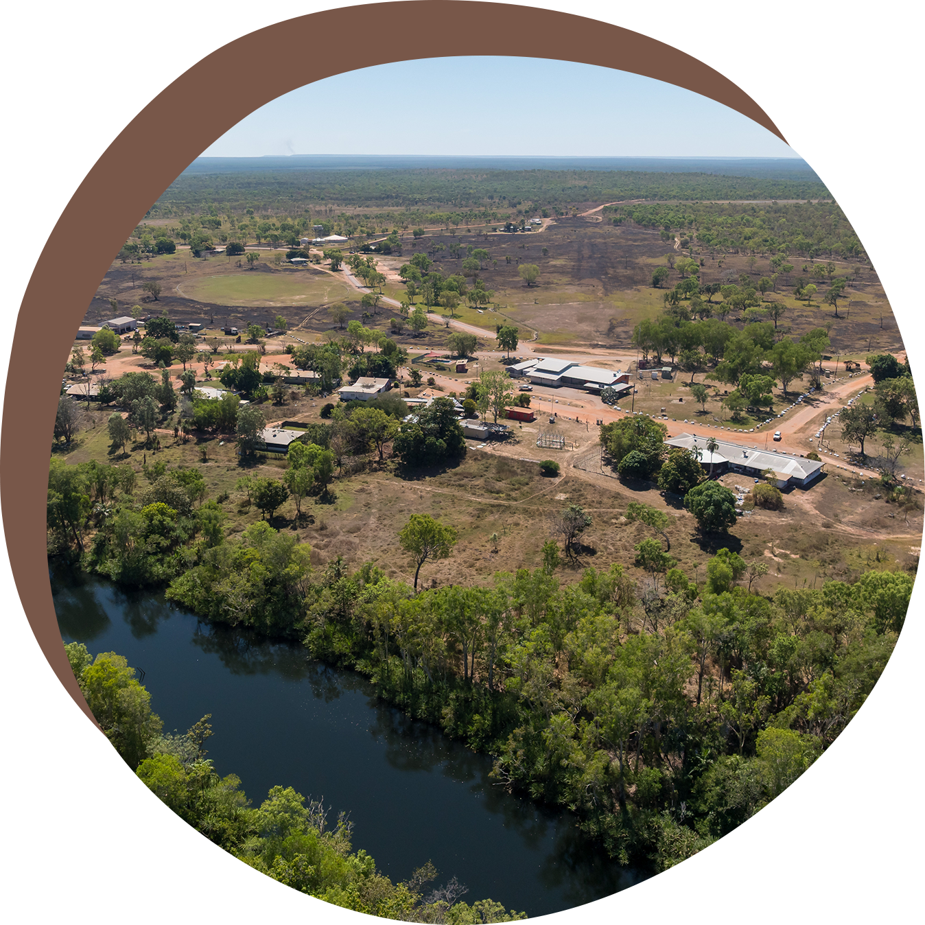 Aerial view of a settlement near a dark river, surrounded by trees and a dry landscape under a blue sky.