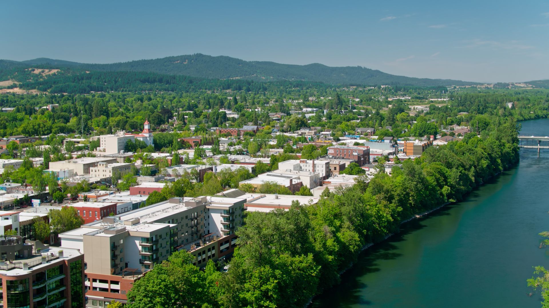 Townscape with buildings, trees, river, and hills under a bright blue sky