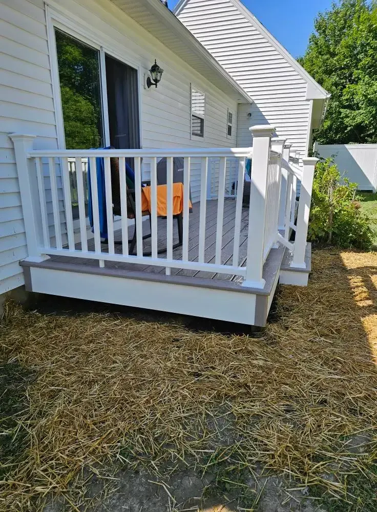 White deck with gray planks and railing, next to a white house. Dry grass in front.