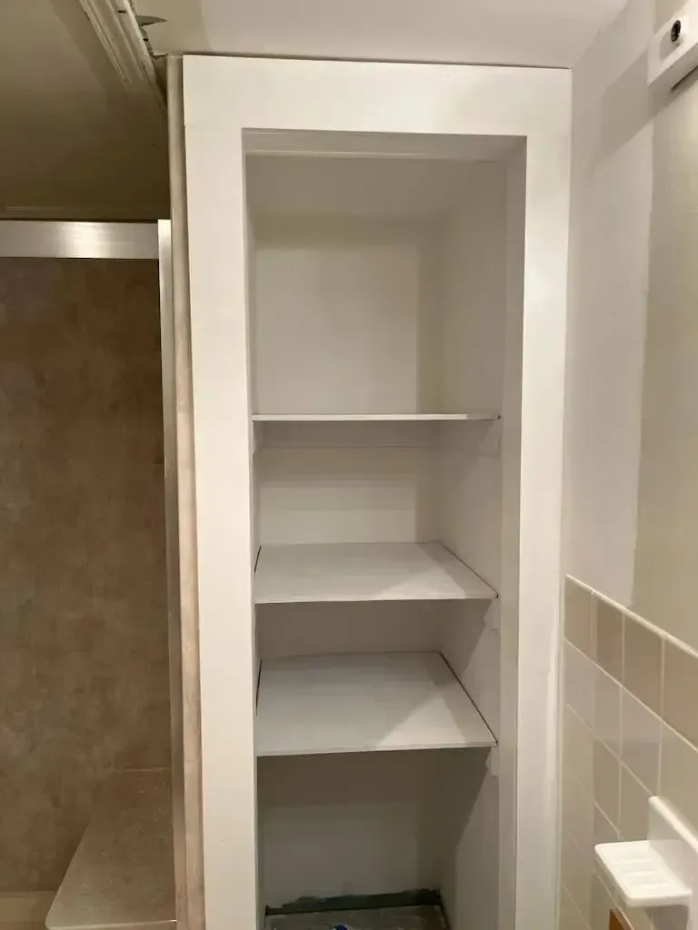 White built-in bathroom storage shelves. Rectangular recess with three shelves; next to a shower and wall.