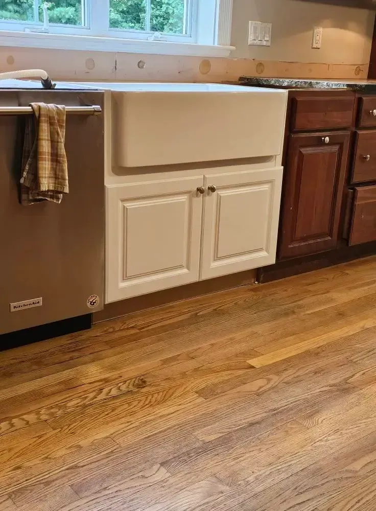 White farmhouse sink with cabinet, adjacent dishwasher, wooden floor and cabinets.
