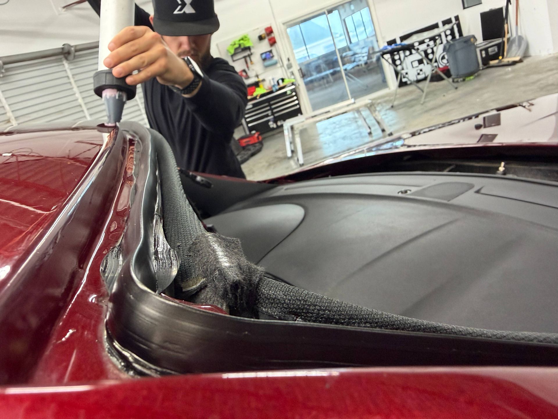 Person cleaning the windshield edge of a maroon car in a garage with a vacuum hose