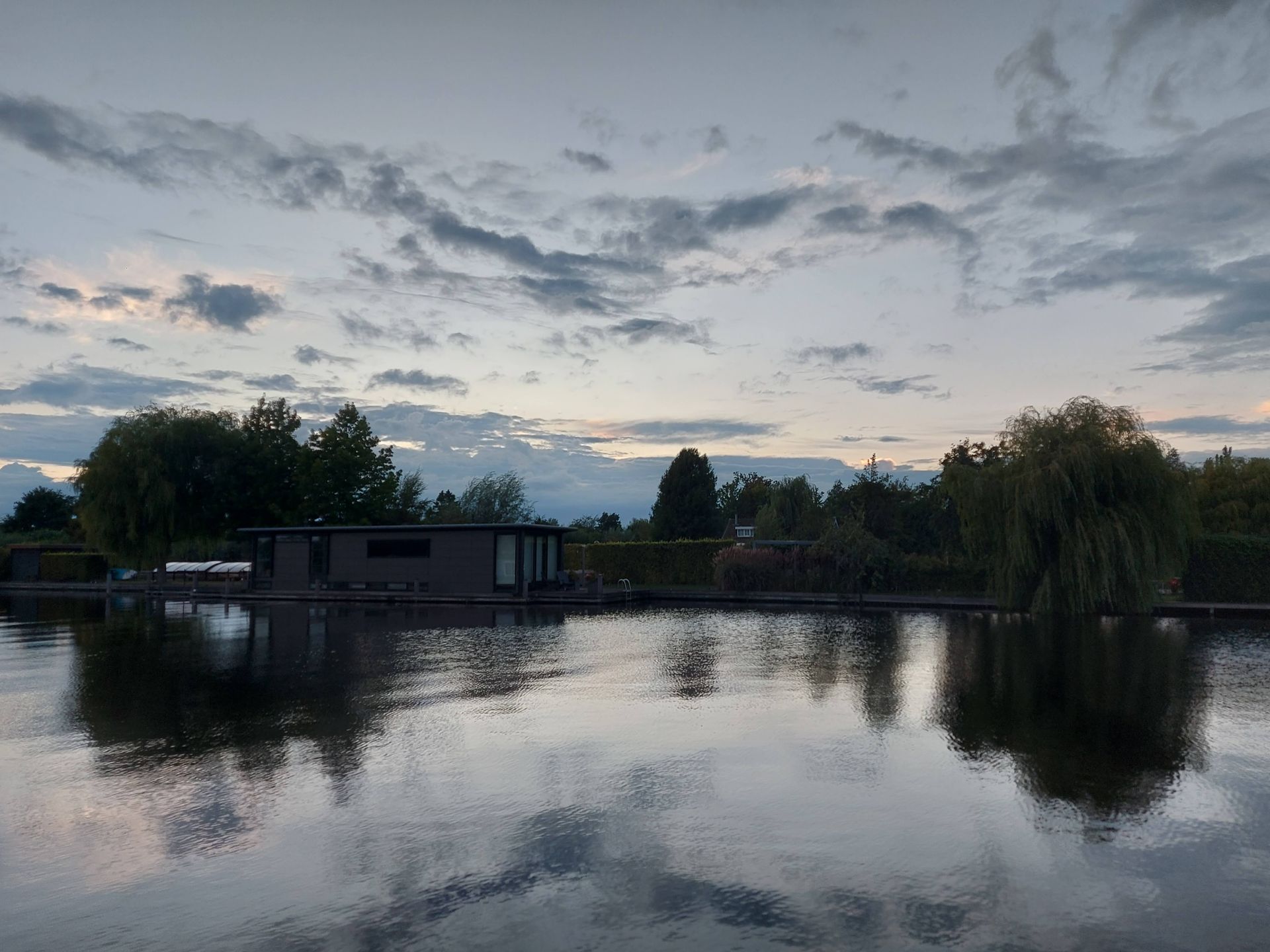 Een modern huis met een plat dak aan de oever van een rivier tijdens een bewolkte zonsondergang, met reflecties op het water.