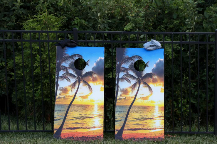 Two cornhole boards with palm trees on them are sitting on the grass next to a fence.
