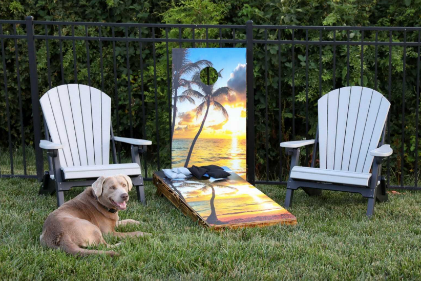 A dog is laying in the grass next to two chairs and a cornhole board.