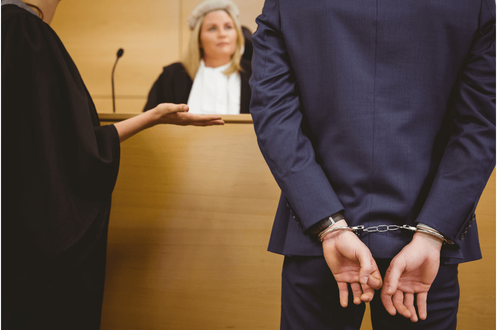 Handcuffed person in a navy suit stands before a judge at a courtroom bench
