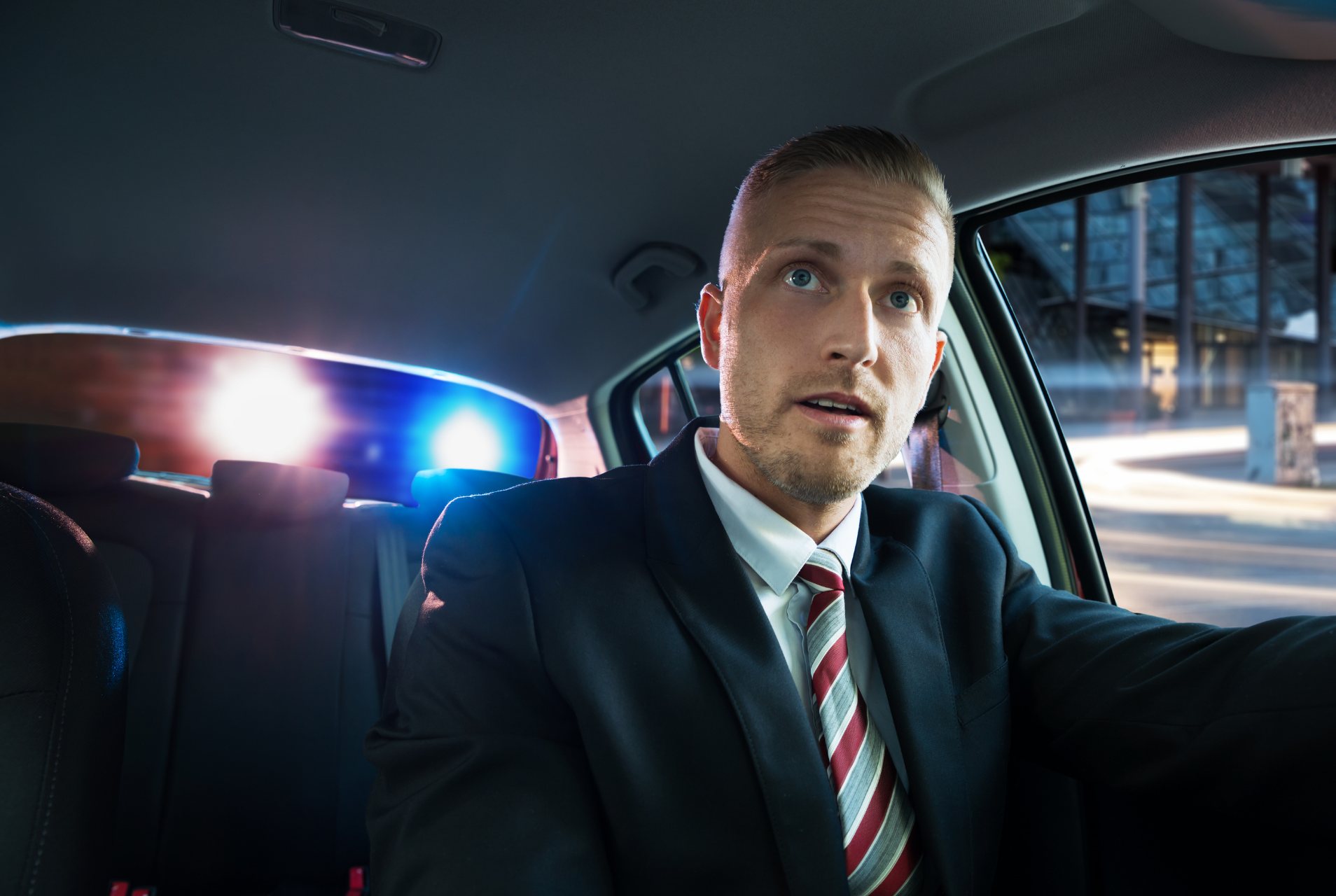 Man in a suit driving a car at night, with flashing police lights behind him