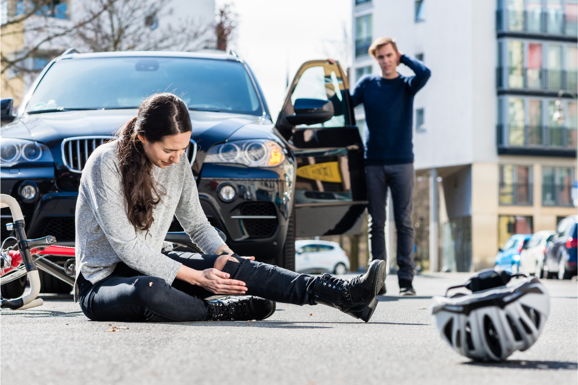Cyclist sitting on pavement by a black car, holding an injured leg, with another person standing nearby.