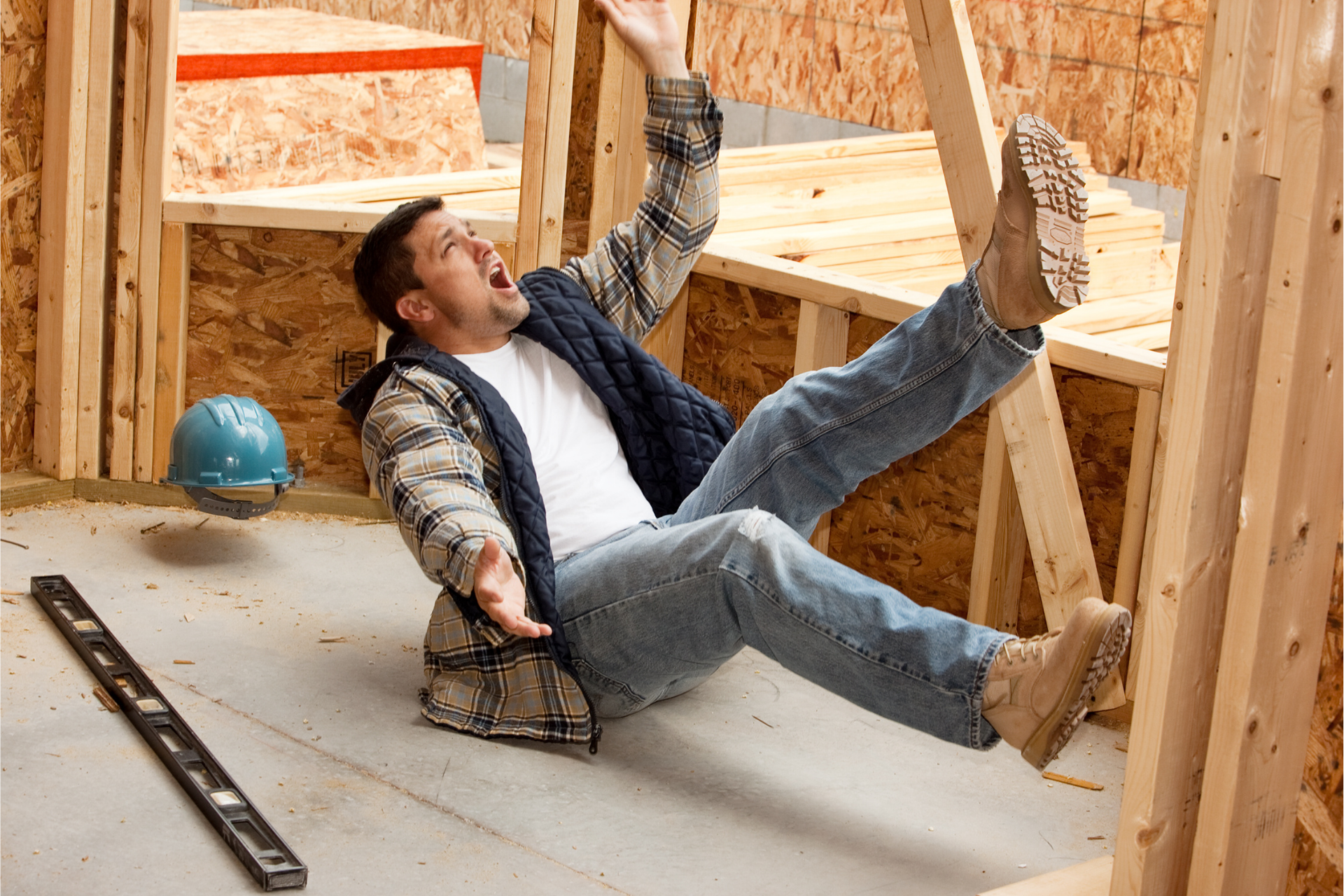Worker falling inside wooden house frame, blue hard hat on floor nearby.