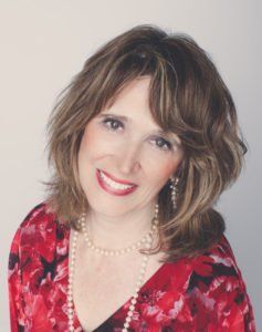 Smiling person in a red floral blouse and pearl necklace against a light background