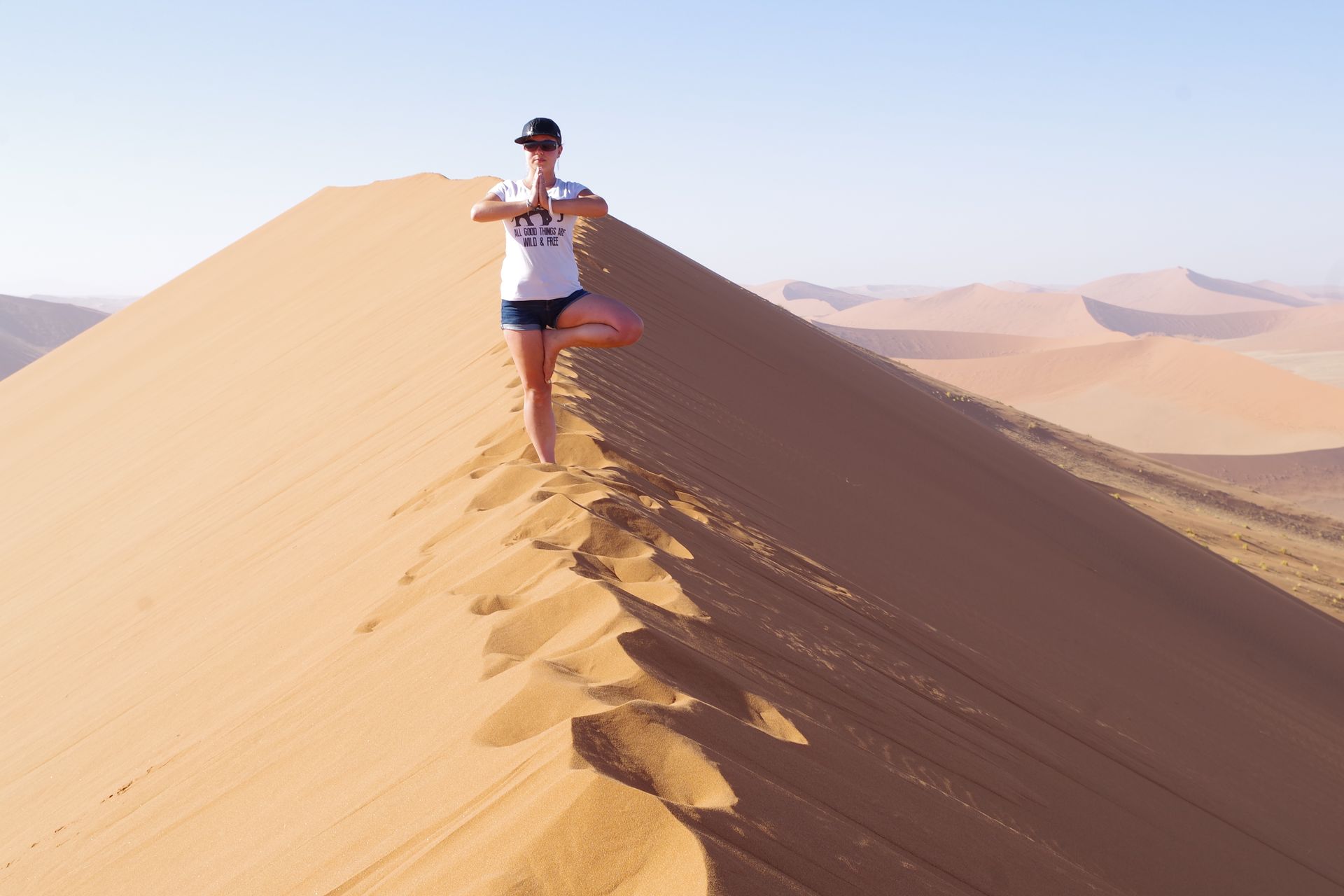 Frau balanciert in einer Baumstellung auf einem Sanddünenkamm in der Wüstenlandschaft.