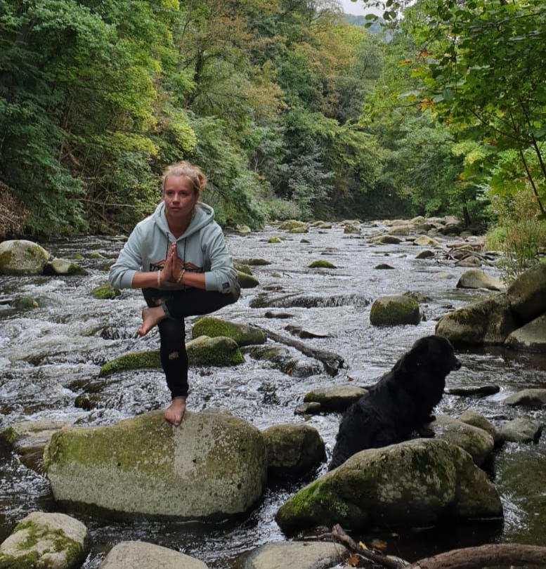 Frau macht Yoga auf einem Flussstein, Hund in der Nähe. Bäume und Wasser im Hintergrund.
