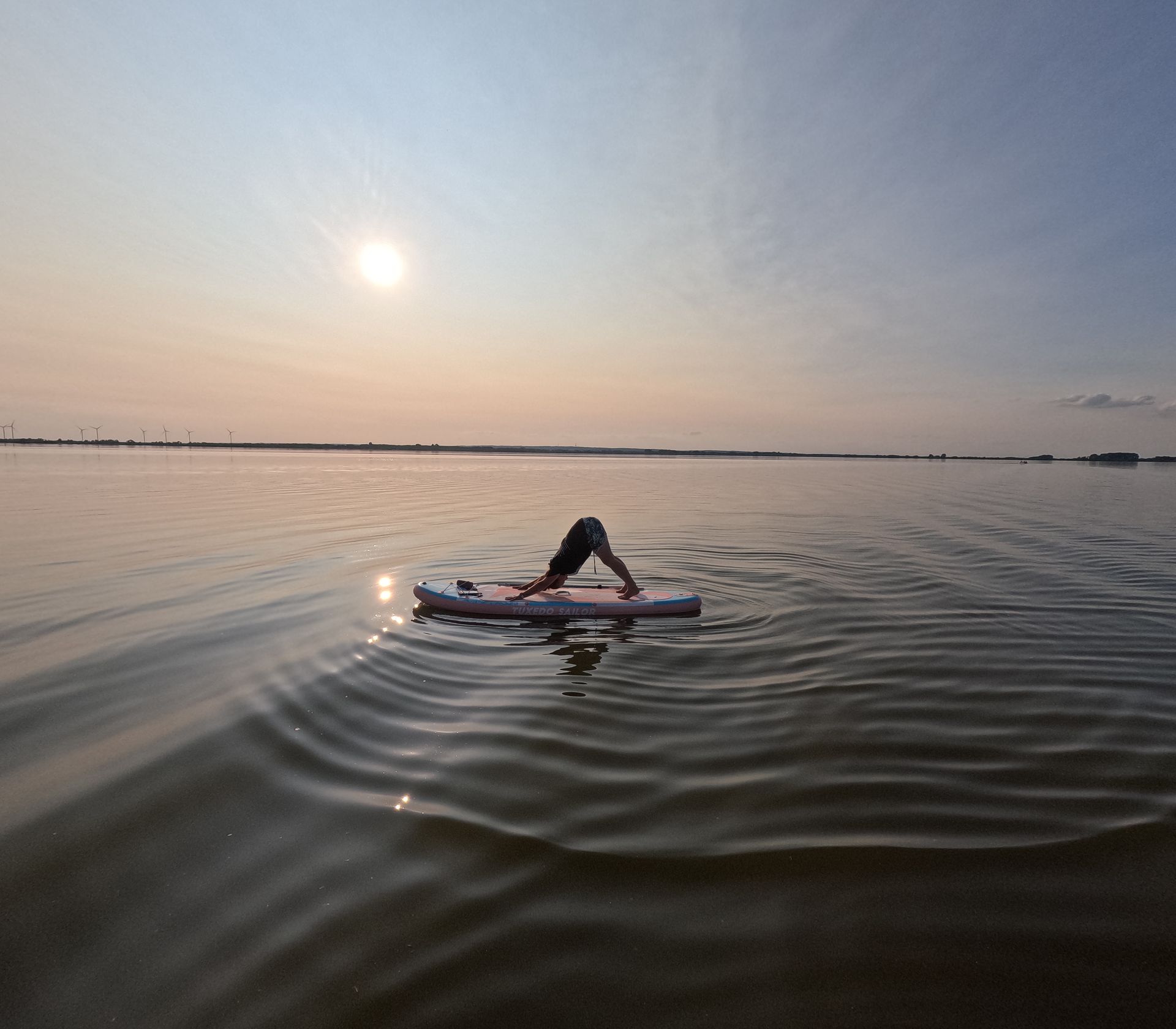 Eine Person auf einem Paddleboard bei Sonnenuntergang im Herabschauenden Hund auf ruhigem Wasser mit leichten Wellen und einer fernen Küste