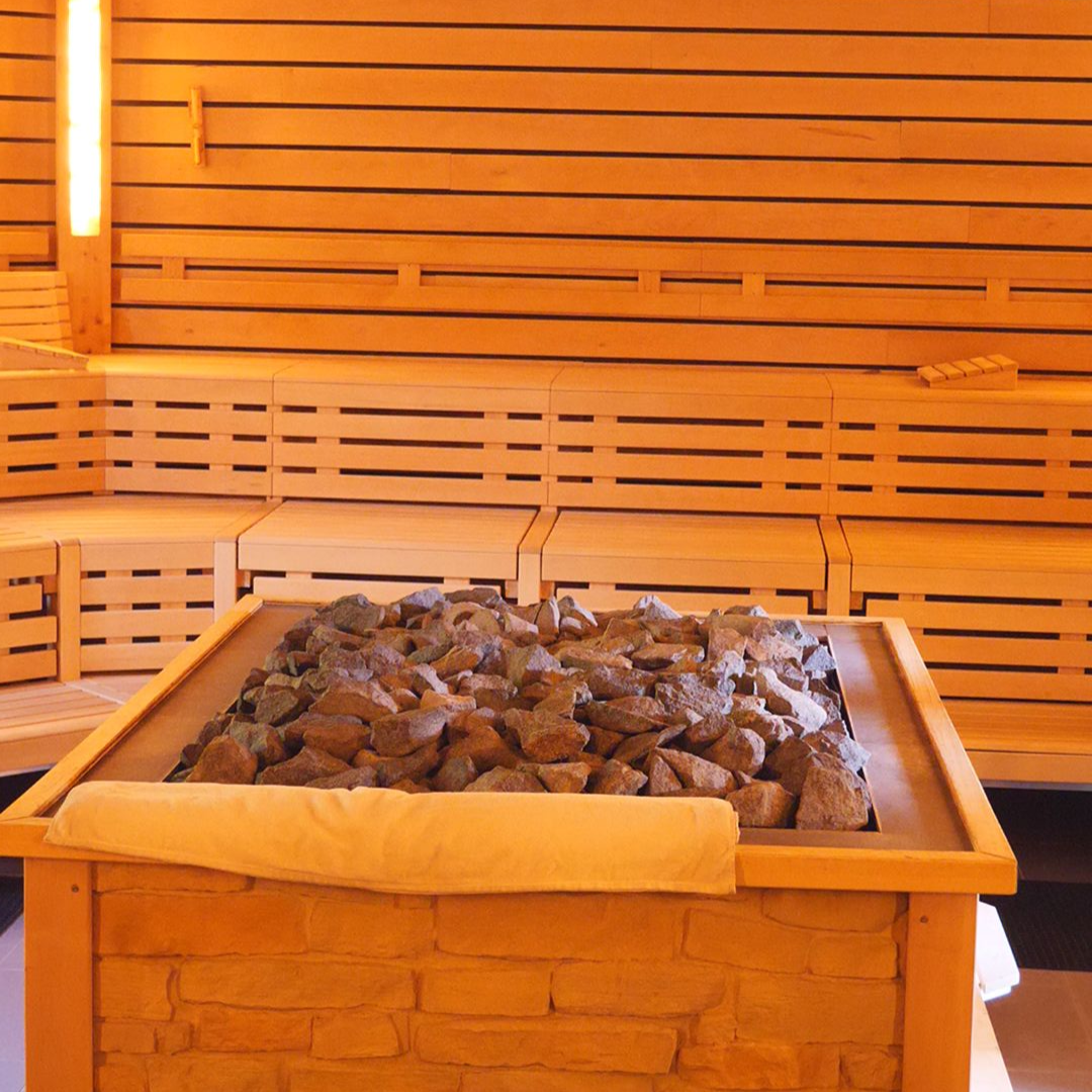 Innenansicht einer Holzsauna mit steinverkleidetem Ofen und mehreren Bänken. Auf dem Ofen liegt ein Handtuch.