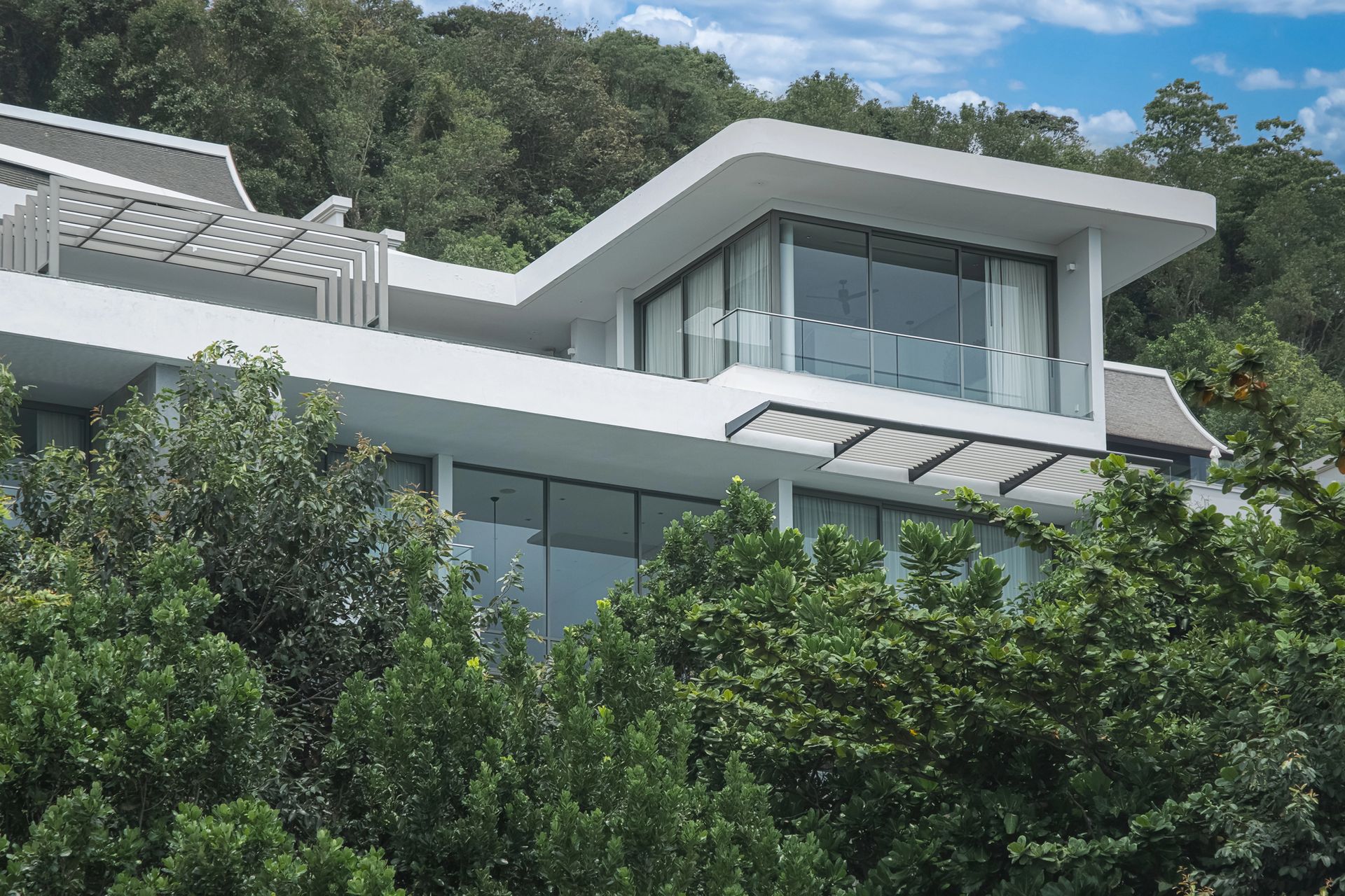 Casa moderna bianca con grandi finestre di vetro, immersa nel verde lussureggiante di una collina.