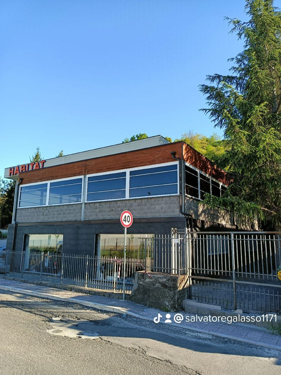 Edificio in mattoni con finestre blu dietro una recinzione nera, sotto un cielo azzurro e limpido.