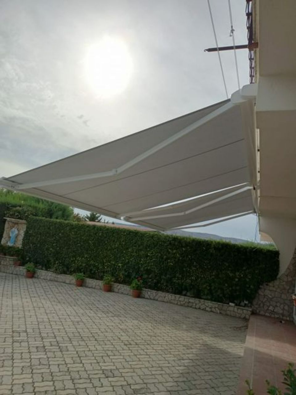 Ampia tenda da sole bianca sopra un patio piastrellato accanto a una siepe, con il sole splendente sopra la testa