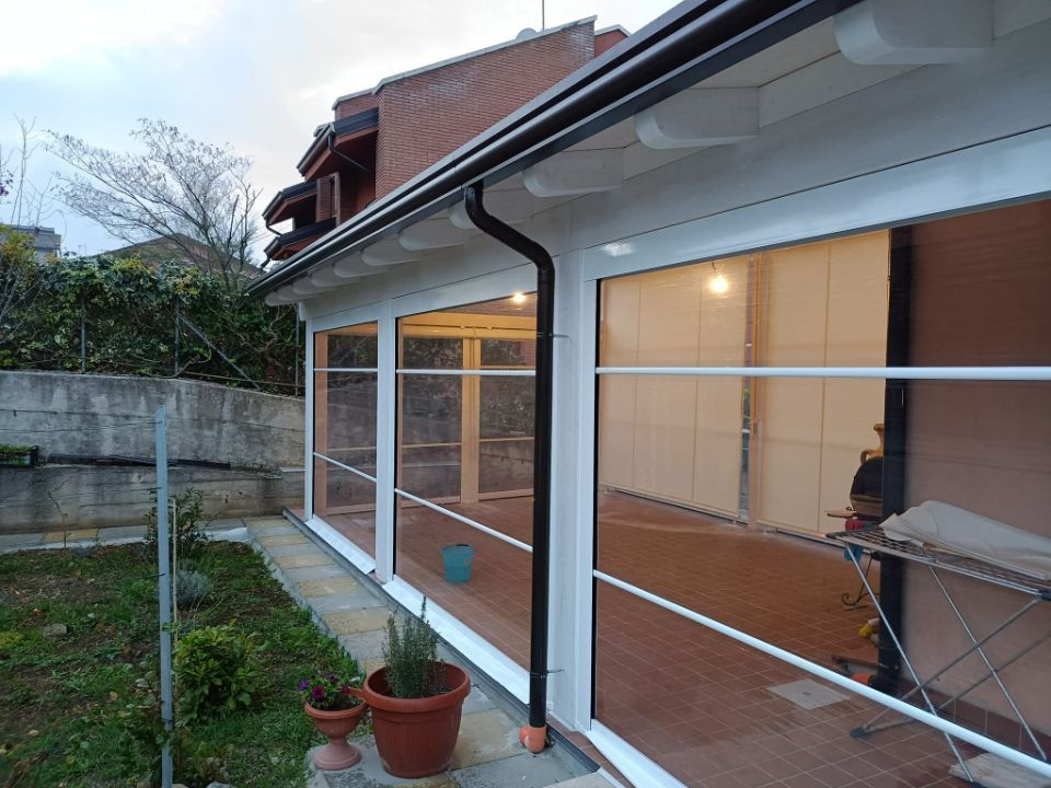 Patio coperto adiacente a una casa, con porte a vetri, piante in vaso e un tavolo da esterno vicino a un prato.