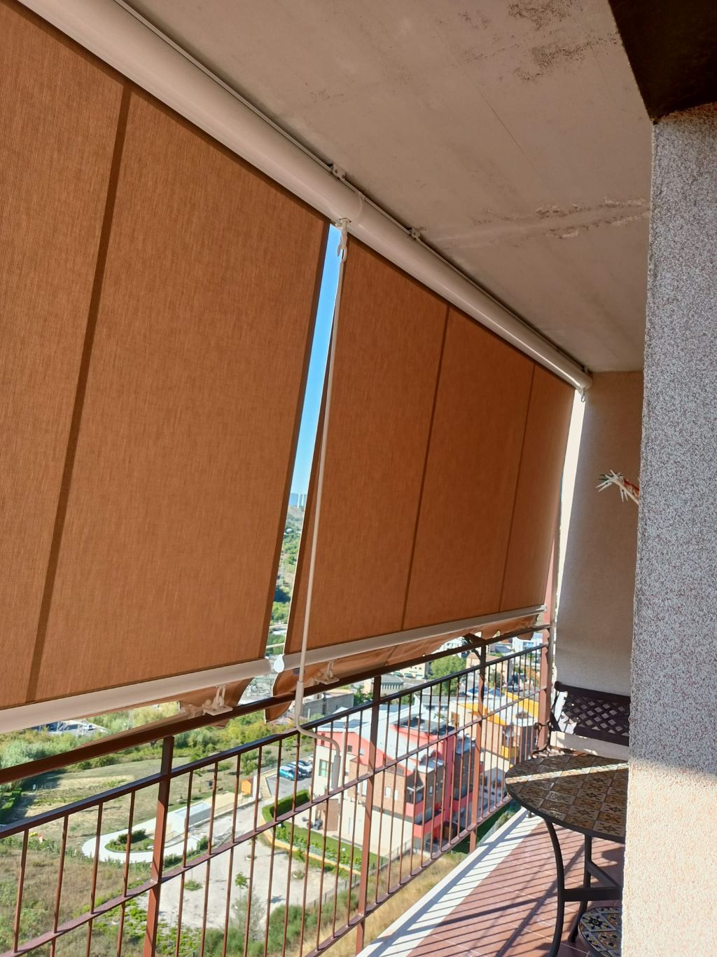 Balcone con tende a rullo color beige, ringhiera in metallo e vista su edifici e vegetazione sottostanti.