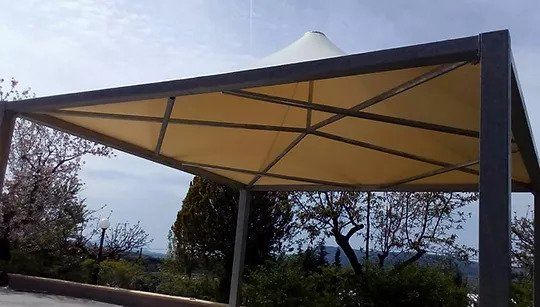 Ampia tettoia beige con struttura metallica, all'aperto, circondata da alberi e con un cielo nuvoloso.