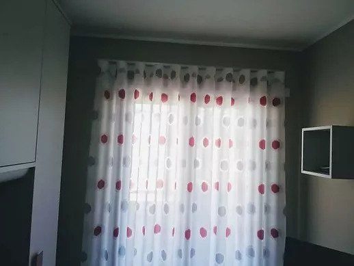 Piccola stanza con una finestra coperta da tende bianche a pois rossi, fiancheggiata da armadi a muro.