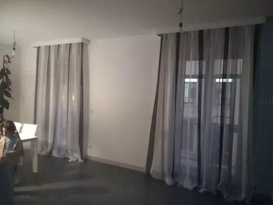 Stanza vuota con tende grigie, una lampada da terra alta e una persona parzialmente visibile sul lato sinistro.