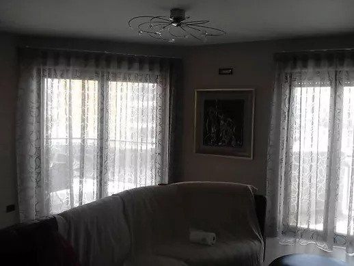 Soggiorno in penombra con finestre dotate di tende trasparenti, ventilatore a soffitto, quadri appesi alle pareti e divano rivestito.