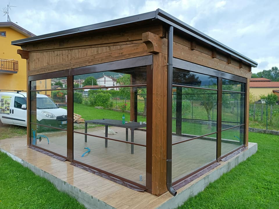 Gazebo in legno con pareti in vetro e lati aperti, posizionato su un prato, con un tavolo all'interno.