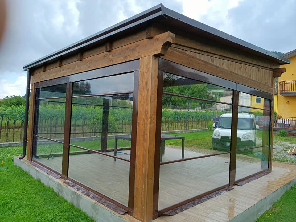 Padiglione in legno simile a un gazebo con grandi finestre in vetro e tetto scuro in un cortile erboso.