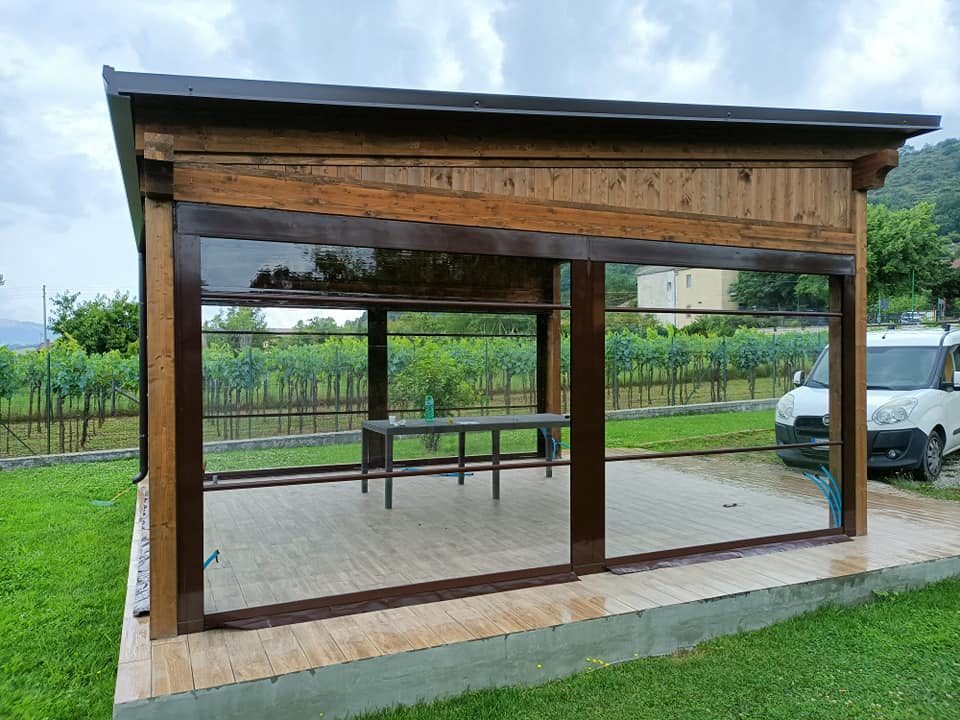 Piccola tettoia in legno aperta sui lati, con tetto di tegole e grandi finestre, situata accanto a una zona pavimentata.