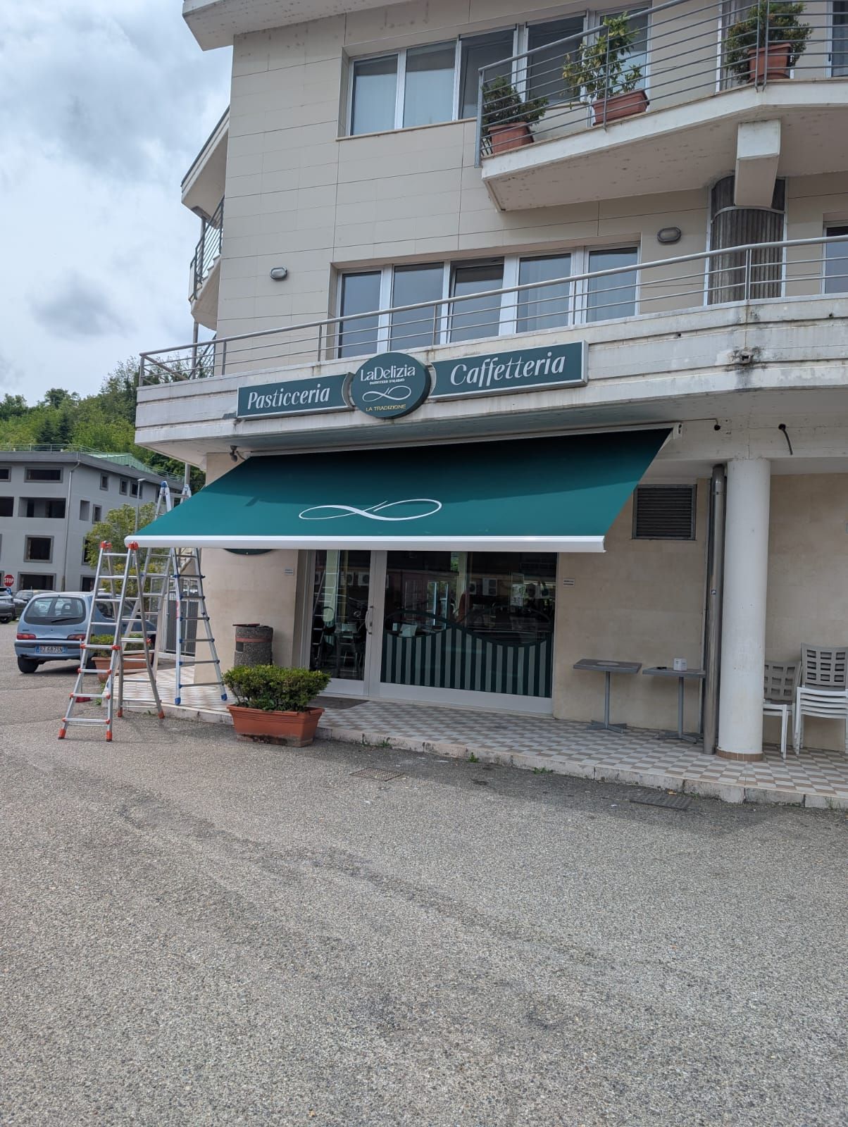 Negozio con tenda da sole verde e balcone su una strada tranquilla, con piante in vaso e un'insegna a cavalletto all'esterno.